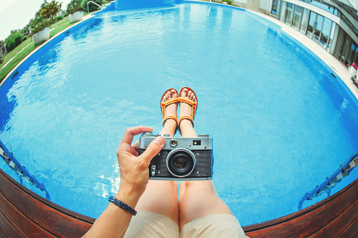 Hand holding old vintage photo camera and make photo of foot sandals against blue water pool background .