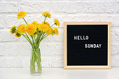 Hello Sunday words on black letter board and bouquet of yellow dandelions flowers on table against white brick wall. Concept Happy Monday. Template for postcard