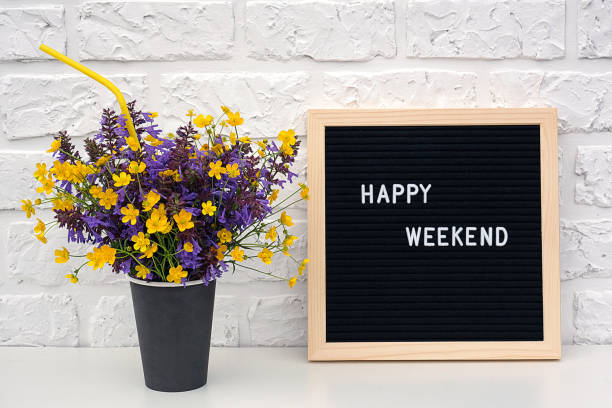 glückliche wochenende worte auf schwarzem brieftafel und strauß von gelben löwenzahn blumen auf dem tisch gegen weiße ziegelwand. konzept happy monday. vorlage für postkarte - today blackboard text letter stock-fotos und bilder