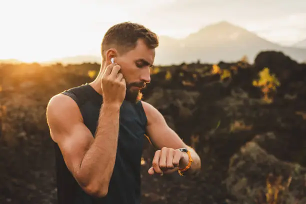 Photo of Male runner synchronizing wireless earphones with smart watch. Preparing for trail running outdoors at sunrise.