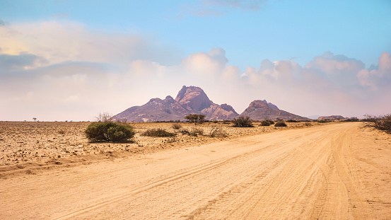 A wide shot of a harsh, desolate African desert landscape, with a long, straight dirt road running toward the dramatic ancient granite peaks of Spitzkoppe in the distance. There are a few bushes and the sky has a few afternoon clouds.