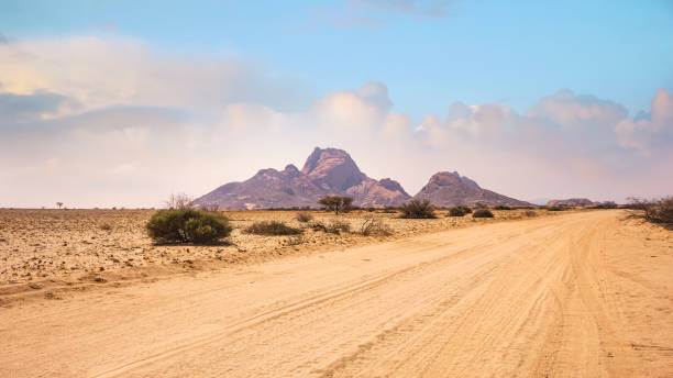 A vast African landscape scene, with a dirt road running through barren flat plains in the Namib Desert toward the huge granite peaks of Spitzkoppe, Namibia. stock photo