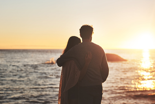 Rearview shot of a happy young couple watching the sun go down at the beach