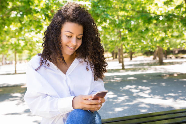 istock Smiling pretty young woman using smartphone on bench in park 1158344622