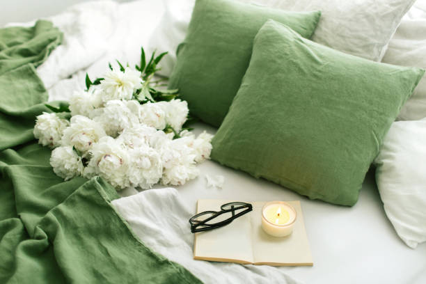Light bedroom and pastel green and gray bedding linen set. Huge white peonies bouquet and reading book with candle. Cozy modern bedroom in fresh spring colours. Morning concept.