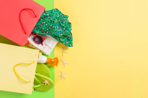Summer concept of colourful shopping bags full of clothes. Gift bags with T-shirt, denim shorts, flip flops and bottle of sunscreen. Summer wishlist concept. Top view copy space on yellow background. concepto veraniego de bolsas de compras coloridas llenas de ropa. bolsas de regalo con camiseta, pantalones cortos de mezclilla, chanclas y botella de protector solar. concepto de lista de deseos de verano. espacio de copia de vista superior sobre fondo am
