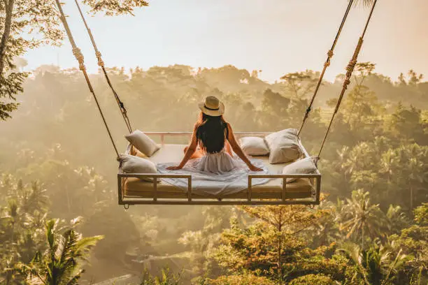 Photo of a young woman sitting on the swing.  Jungle Bed hanging over the tropical forest with Caucasian female resting while looking at the view, Bali, Indonesia. Rear view of a female sitting and enjoying the view.