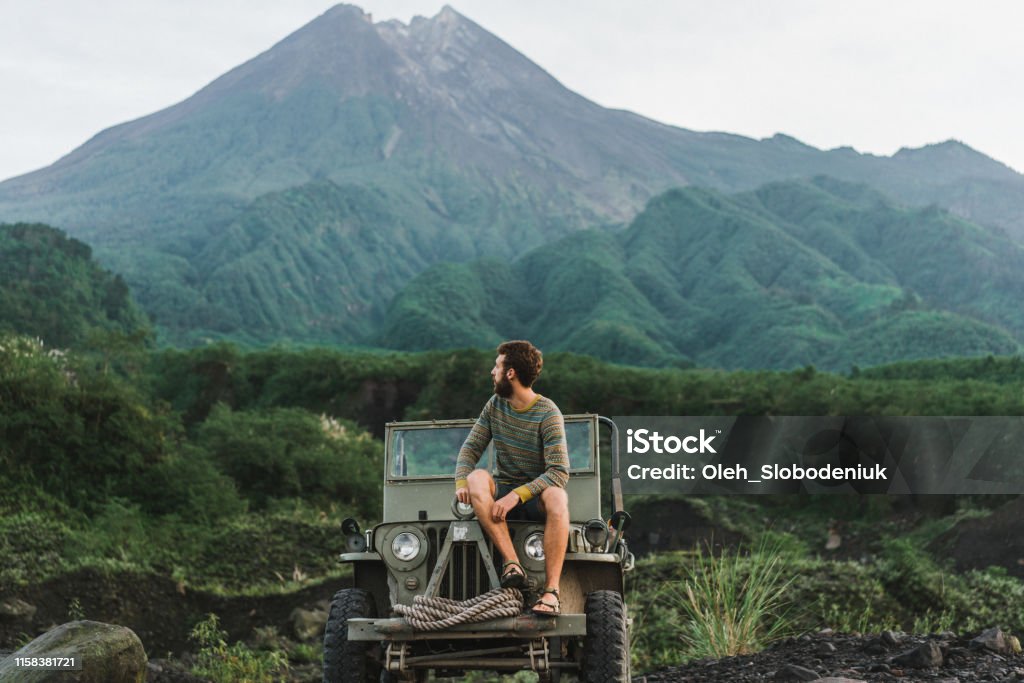 Man sitting on old fashioned SUV on the background of Merapi volcano Scenic view of young Caucasian man sitting on old fashioned SUV on the background of Merapi volcano Rainforest Stock Photo