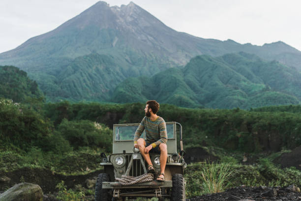Man sitting on old fashioned SUV on the background of Merapi volcano Scenic view of young Caucasian man sitting on old fashioned SUV on the background of Merapi volcano bromo tengger semeru national park stock pictures, royalty-free photos & images