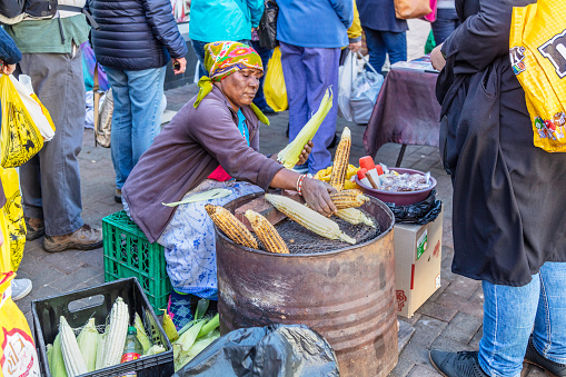 Senior lady selling roasted mielies or corn cobs, on the barbeque grill, a market vendor  in Fordsburg, Johannesburg.\nFordsburg is an old historical area where mainly indian, turkish, pakistani and other east asian cultures coexist with restaurants and spice dealers.