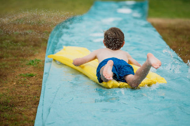A front view shot of a young boy sliding down a water slide on his back in a garden, he is having a fun time.