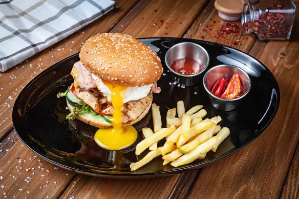 Fresh burger with chicken egg, lettuce and sauce on a black plate with french fries. American fast food. Chickenburger with copy space on wooden background. Close up, selective focus. food. Grill menu