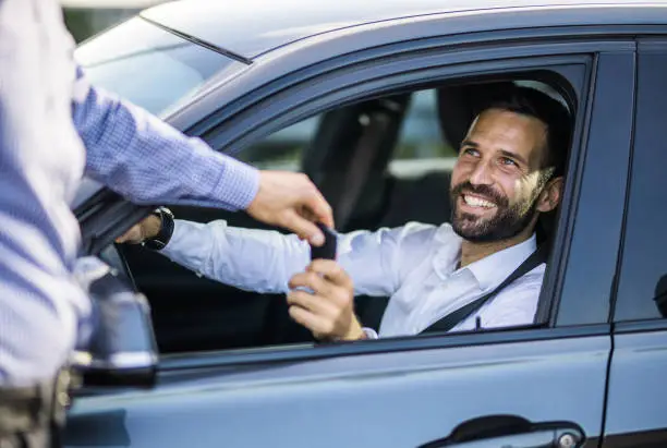 Young happy businessman sitting inside of his new car and taking car keys from a sales person.