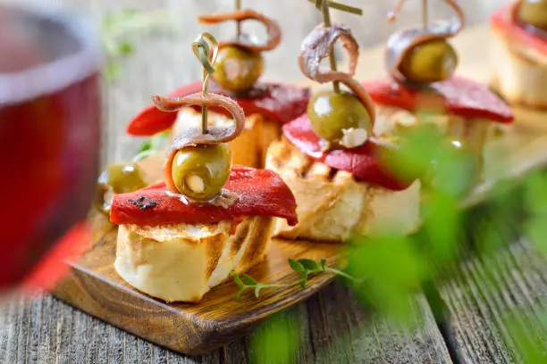 Spanish pinchos: Skewer snacks made of grilled bread with roasted and pickled peppers, olives stuffed with cream cheese and parmesan and on top delicious anchovy fillets, served with a glass of red wine