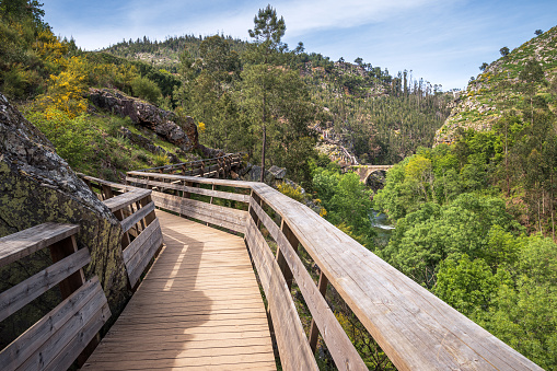 Beginning of the Passadiços do Paiva along the Areinho river beach, seen with part of the wooden walkway in the foreground and in the background the Garganta do Paiva with a stone bridge in Arouca, Portugal.