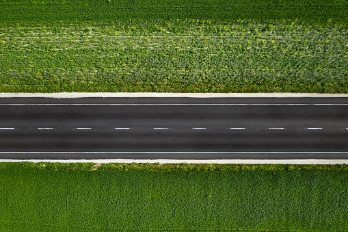 Straight empty road seen from above, surrounded by green grass and meadows, crossing the image horizontally with two lanes. Aerial photography shot from drone.
