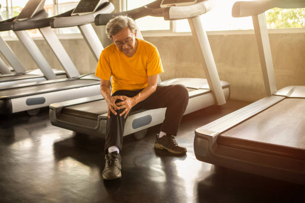 Senior asian sport man injury knee pain  sitting on treadmill in fitness gym . aged  suffering from Arthritis elder male exercising , workout, training ,healthy ,Retirement ,older, with copy space stock photo