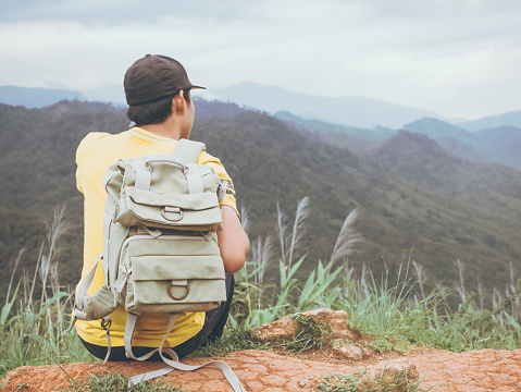 camping and travel hiking concept from backside of hipster young man with backpack sit see to valley landscape view on peak mountain in tropical rainforest with happiness and freshness filling.