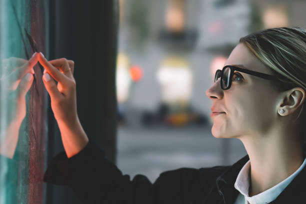 istock Smiling female standing at big display with advanced digital technology. Young woman touching with finger sensitive screen of interactive kiosk for find information while standing on street in evening 1158458512