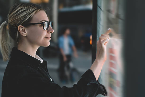 istock Smiling female standing at big display with advanced digital technology. Young woman touching with finger sensitive screen of interactive kiosk for find information while standing on street in evening 1158458526