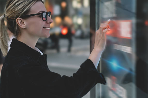 istock Smiling female standing at big display with advanced digital technology. Young woman touching with finger sensitive screen of interactive kiosk for find information while standing on street in evening 1158458533