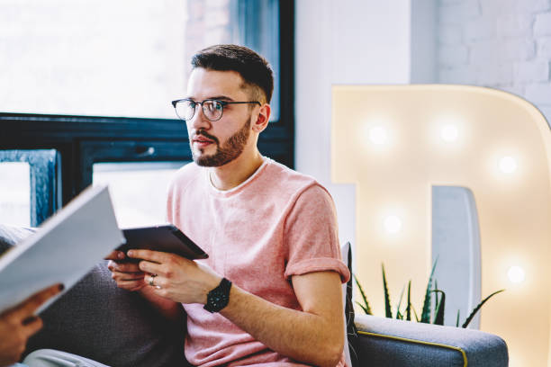 istock Bearded young man in eyeglasses for vision correction updating software on modern tablet device using wireless internet connection in modern apartment.Male blogger downloading app on touch pad at home 1158471658