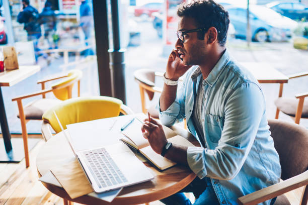 istock Pondering professional freelancer in cool spectacles having call with customer using modern smartphone and internet connection.Pensive hindu male spending time in coffee shop and talking on phone 1158473261