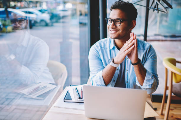 istock Handsome positive smart male entrepreneur in spectacles enjoying time while pondering about new ideas for business startup.Successful man freelancer looking outside while sitting at table with laptop 1158473285