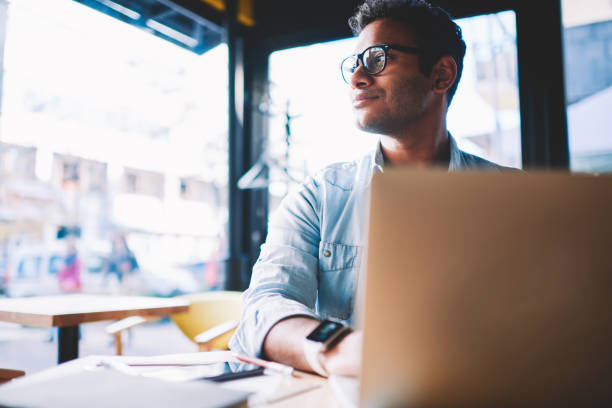 istock Pondering hindu young man looking away while working remotely at modern laptop with wireless 4G internet.Contemplative hipster student thinking on creative ideas for coursework sitting in coworking 1158473657
