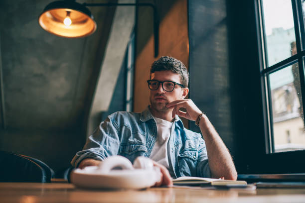 istock Thoughtful young hipster guy in eyewear pondering on organisation plan sitting at desktop indoors,contemplative caucasian male student spending time for learning at university campus thinking on essay 1158475787