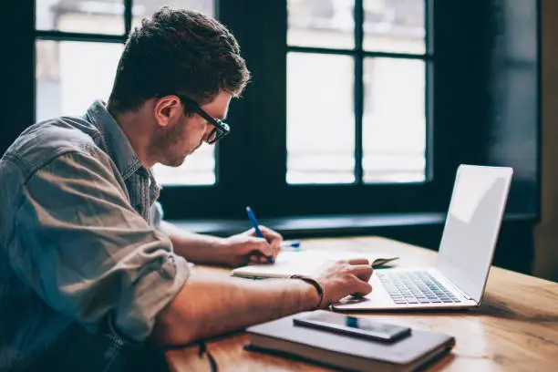 Photo of Young millennial male student searching information for homework using laptop and wifi connection indoors, man writing article in textbook for add interesting content on own website or web page