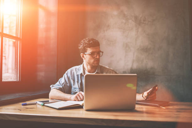 istock Handsome male it professional checking time on smartphone device and updating notification indoors, casual hipster guy sitting in front of laptop and dialing number on cellular phone via app 1158475845