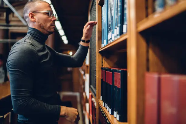 Serious male journalist standing near bookshelves picking literature for researching information, concentrated smart historian selecting books in public library working with periodical documents