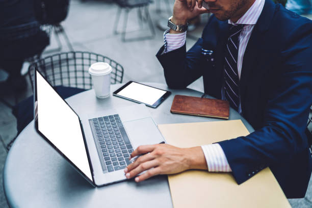 istock Cropped view of professional businessman sitting at table front open laptop computer with blank mock up screen, close up entrepreneur in suit working remote on netbook and talking on cellphone 1158476110