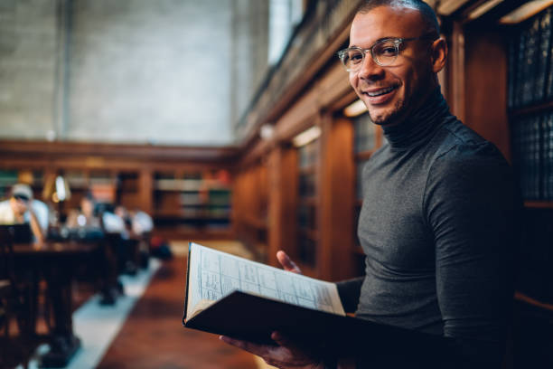 istock Half-length portrait of smiling young academic finding out interesting information during research, cheerful journalist in eyeglasses standing in public library increasing knowledge about literature 1158476169