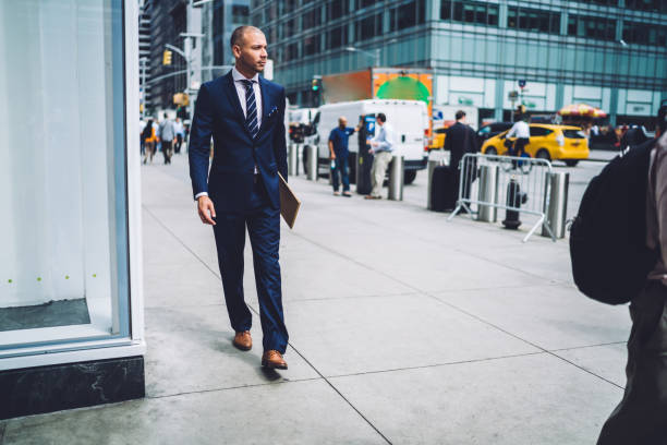 istock Confident young man lawyer dressed in formal wear holding folder in hand and strolling on street in downtown.Successful businessman in elegant suit walking in urban setting of modern city 1158476313
