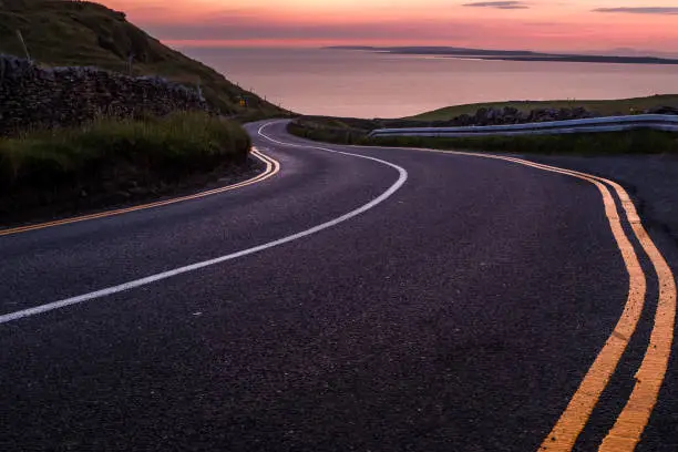 Photo of Looking down a sweeping road near the Cliffs of Moher to beautiful pink sky sunset over the island of Inisheer.