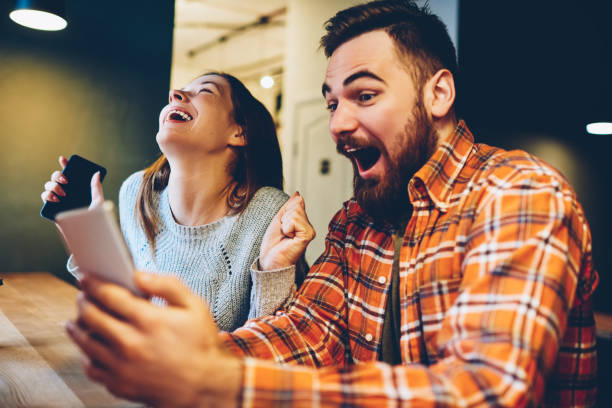 os hipsters masculinos e fêmeas excited regozijam-se em ganhar uma loteria do internet fizeram apostas no web site no smartphone moderno. pares felizes no amor que comemora a vitória em competições em linha que apreciam o sucesso - jogos de azar - fotografias e filmes do acervo