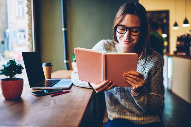 istock Happy skilled student in eyewear reading interesting information in textbook preparing for studying seminar in coworking space,Smiling young woman in casual apparel enjoying bestseller in leisure time 1158477968