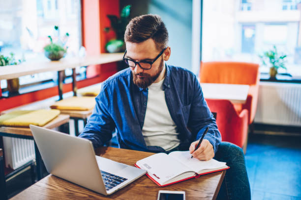 istock Smart bearded student searching information on websites and making notes in notepad sitting in stylish coworking space and using 4g internet connection.Hipster blogger planning working process 1158478001
