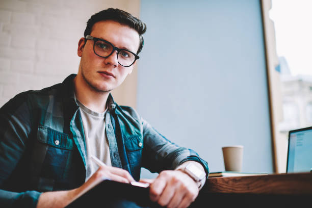 istock Portrait of serious hipster guy in spectacles for vision correction making notes during distance job process, professional male freelance writing articles for publication looking at camera in cafe 1158478825