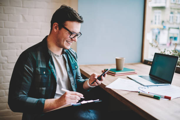 Cheerful young man laughing on funny video browsed on smartphone while working remotely in cafe ,smiling hipster guy satisfied with getting good news on email checking notifications on telephone joven alegre riendo en video divertido navegado en el teléfono inteligente mientras trabajaba de forma remota en el café, sonriendo hipster chico satisfecho con conseguir buenas noticias en las notificaciones de comprobación de correo electrónico en el