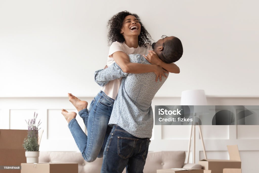 African husband lifting happy wife celebrating moving day with boxes African husband lifting happy wife laughing celebrating moving day with boxes, excited first time home buyers renters owners having fun enjoy relocation into own new flat house, mortgage investment Couple - Relationship Stock Photo