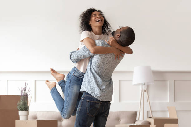 African husband lifting happy wife celebrating moving day with boxes African husband lifting happy wife laughing celebrating moving day with boxes, excited first time home buyers renters owners having fun enjoy relocation into own new flat house, mortgage investment reform photos stock pictures, royalty-free photos & images