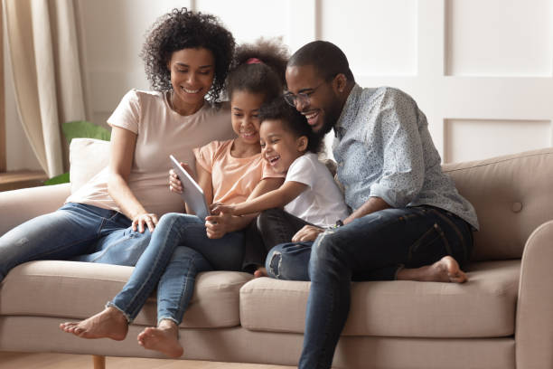 Happy black parents and children using digital tablet on sofa Happy african family having fun with device at home, black parents and little children using digital tablet looking at screen sit on sofa together, cute kids hold computer laugh watch cartoons online african american children stock pictures, royalty-free photos & images
