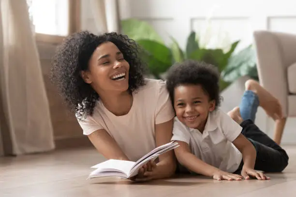 Photo of African mom reading book laughing with child son at home