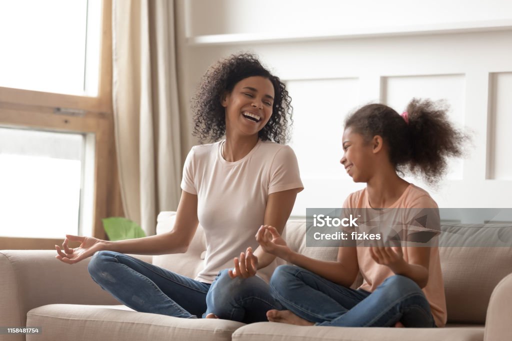 Healthy happy african mother teaching yoga happy kid daughter laughing Healthy happy african american mother teaching yoga happy kid child daughter laughing having fun doing exercises enjoy meditating relaxing sitting in lotus position on sofa in living room at home Family Stock Photo