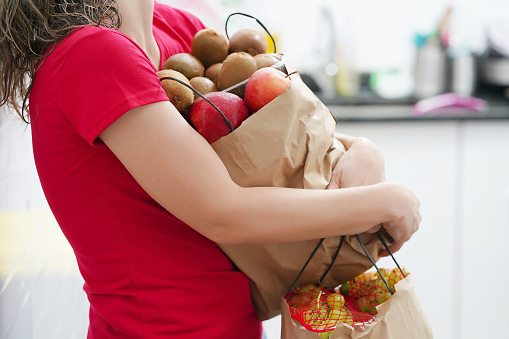 Woman holding a paper bag with food. Housewife with food packages after a supermarket in the hallway of her apartment. Housewife with a paper bag full of fruits in the kitchen.