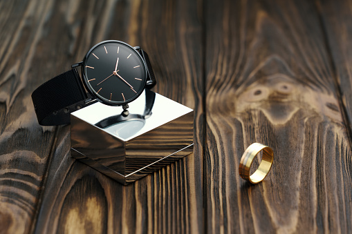 Black modern wrist watch on chrome cube with golden ring on wooden desk