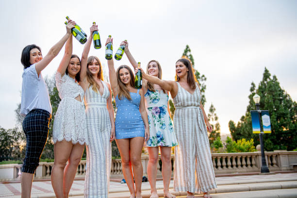 Six Female College Students Celebrate Graduation With Champagne Bottles Raised High In The Air Six college roommates/sorority sisters celebrate graduation by popping champagne and toasting the bottles together with arms extended above their heads. sorority photos stock pictures, royalty-free photos & images